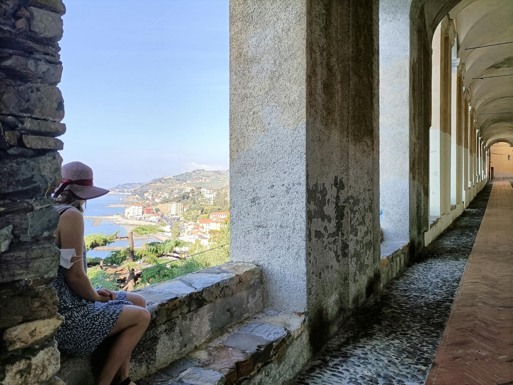 A woman with a hat sits on a ledge overlooking the scenic coastal city of Imperia, Italy.