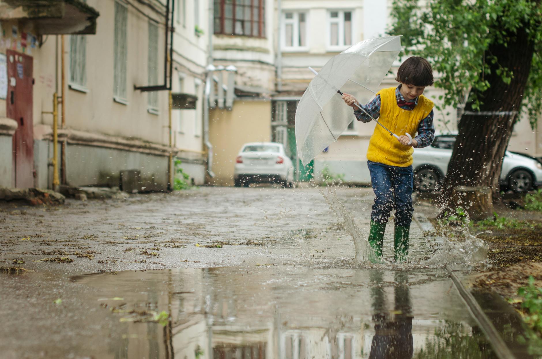 A child joyfully splashes in puddles on a rainy day, holding an umbrella in a quaint neighborhood street.