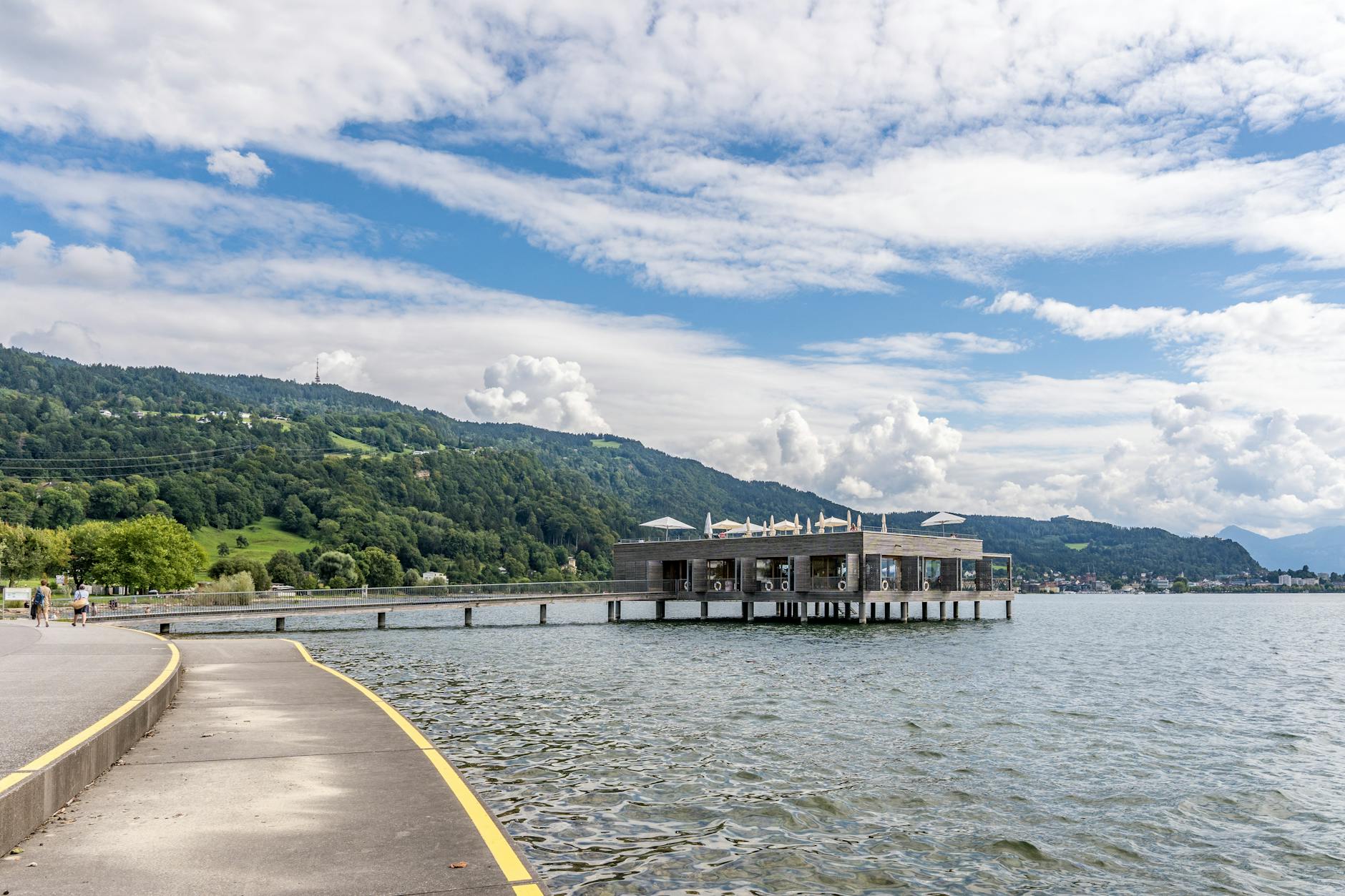 A serene lake with a modern pier, hills, and promenade under a vibrant sky.