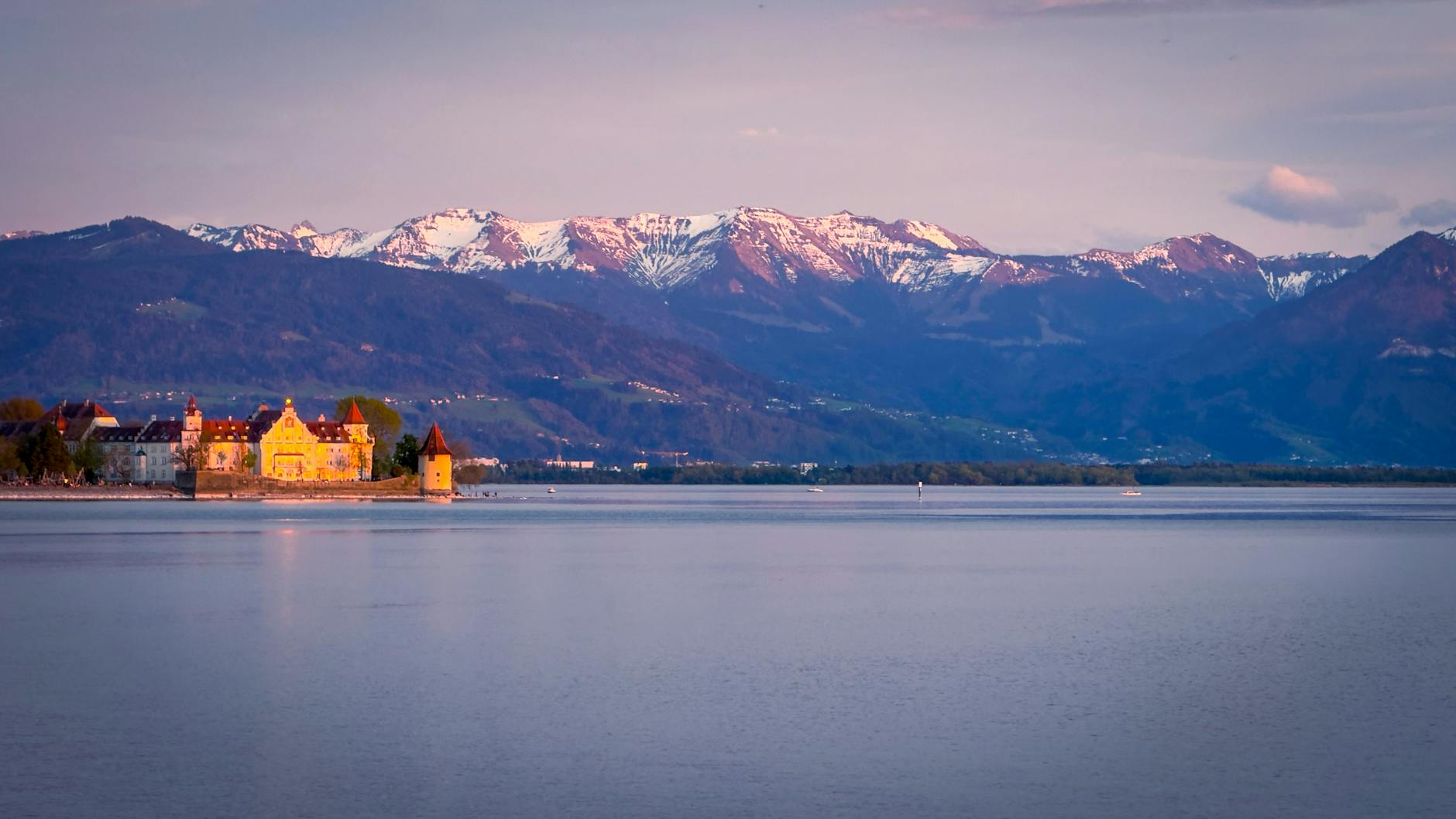 Sunset view of a castle by a tranquil Alpine lake with majestic mountains in the background.
