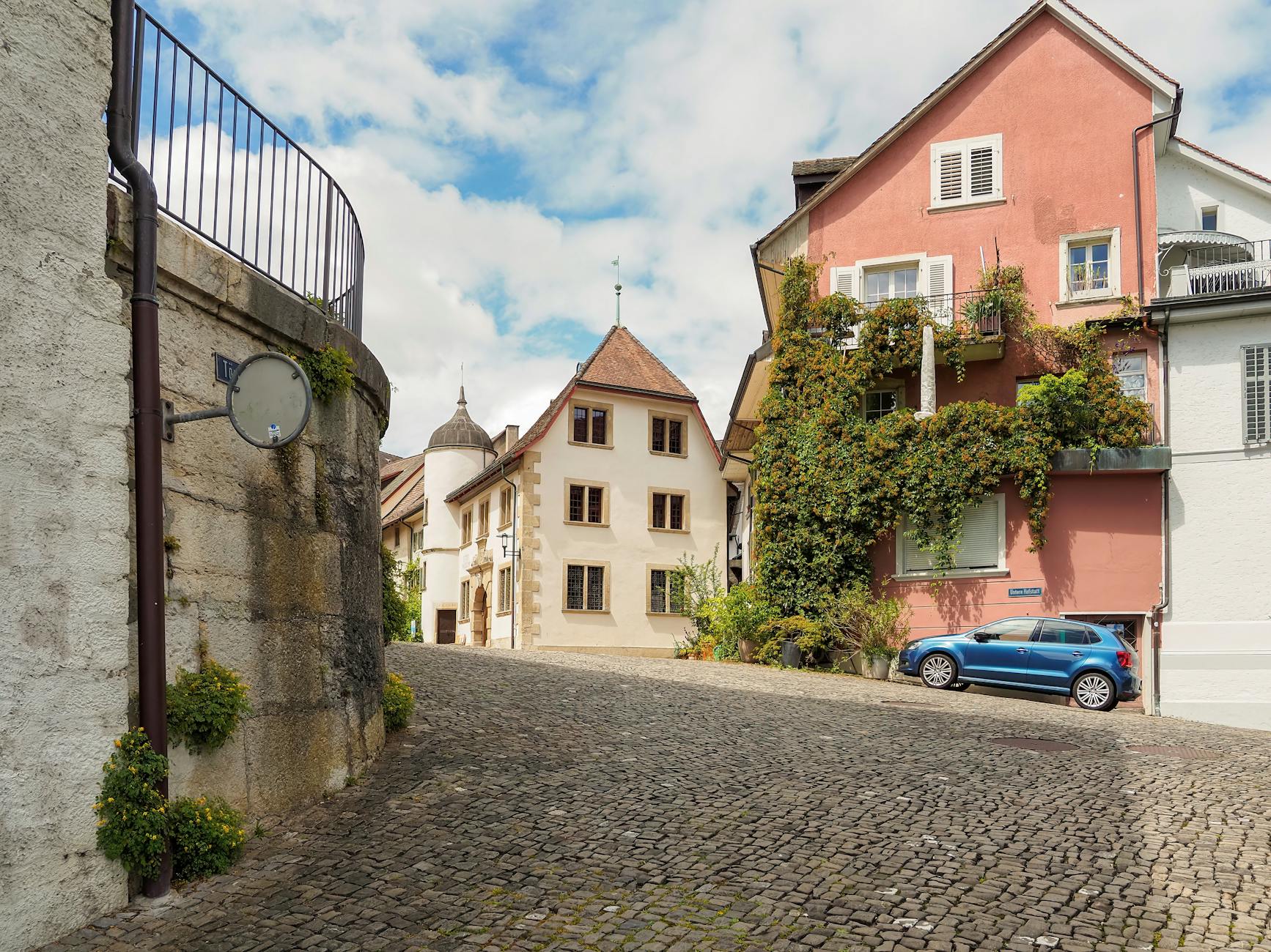 Cobblestone street in a quaint European town with ivy-covered houses and a parked car on a sunny day.
