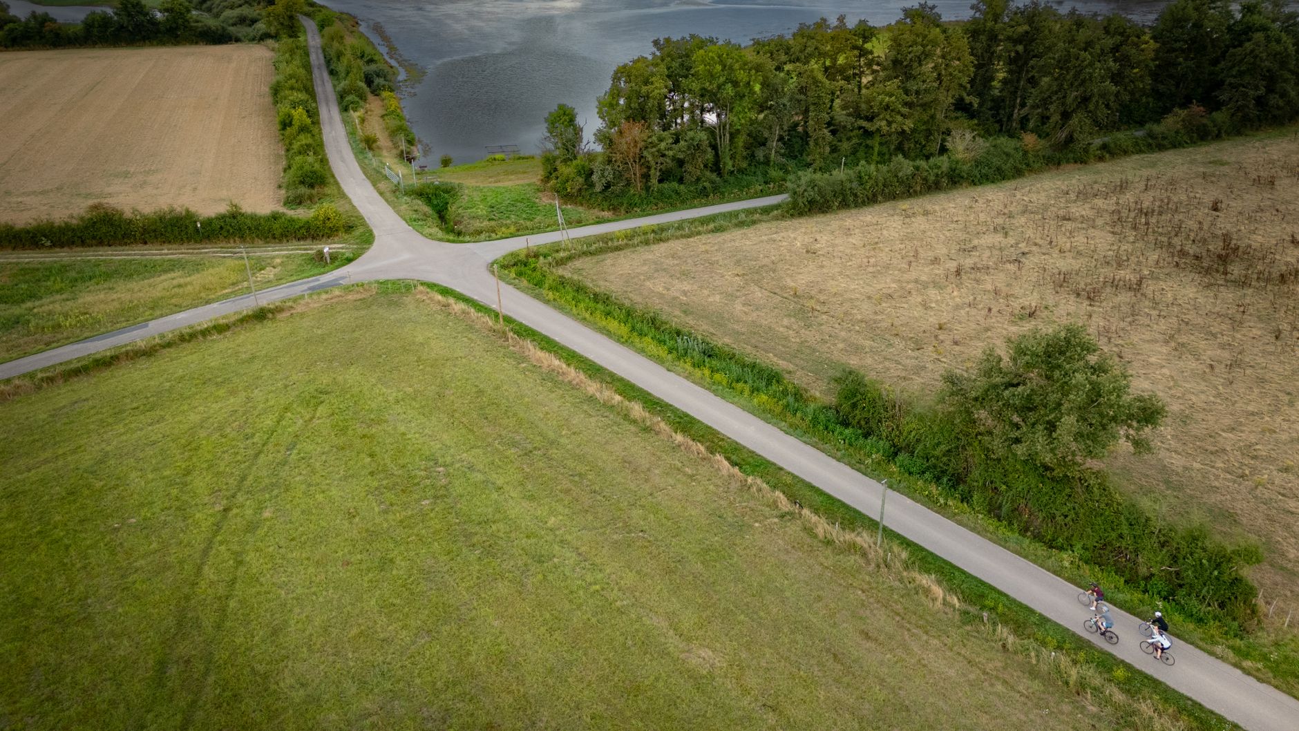 Aerial view of cyclists on a rural road near a tranquil lake intersection, surrounded by fields.